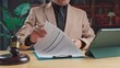 © Mdisk - Mature lawyer in formal suit reviewing legal contract papers near wooden gavel digital tablet inside courtroom office workspace.