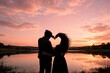 © ArtAvion - VALENTINE'S DESIGN: Romantic silhouette of a young couple standing by a quiet lake at golden hour, framing a heart symbol with their hands against a colorful pink and orange sunset