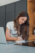 © Daniel - Focused young woman writing in a notebook beside her laptop in a modern kitchen workspace, concentrating on planning, studying and remote work with a calm, productive and casual atmosphere.