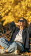 © pattozher - Autumn Serenity: A woman in casual autumn attire, wearing sunglasses and a blazer, relaxes on a park bench amidst a shower of golden leaves, embodying peace and contentment.