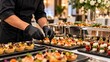 © aisyah karim - Catering professional in black uniform and gloves arranging gourmet appetizers and canapés on a buffet table at an event with chafing dishes in background