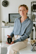 © AnnaStills - Vertical portrait of middle aged Caucasian businesswoman sitting on desk holding cup in office. Image suitable for corporate communication and professional leadership campaigns
