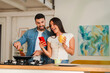 © Jose Calsina - A happy couple cooks side by side at a kitchen island, one holding a red phone while the other stirs a frying pan and wears a white top. Nearby a mug sits on the counter as they smile and share a
