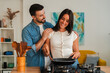 © Jose Calsina - A warm scene shows a couple enjoying food prep at a stove, one stirs a skillet while the other smiles and assists. Wooden utensils stand in a holder, emphasizing teamwork and shared cooking