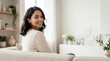© Alva - A beautiful young woman smiling and looking back while sitting on a cozy sofa at home.