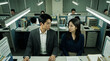 © Bboy - Asian Colleagues Sitting Side by Side at Cubicle Desks Gazing Tensely Under Lights