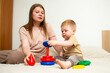 © Krievietka - Mother and two year old son stacking colorful pyramid rings on bed. Parent child play teaches size order and logic. Fine motor skills development through classic toy