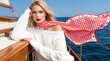 © FRANK - An elegant woman in autumn attire enjoys a carefree boat trip at sea, wearing a white boat-neck sweater paired with a red-and-white checkered scarf featuring a cherry pattern.