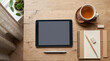 © CLOSE-UP - Digital tablet with a blank screen on a wooden desk with a cup of tea, notepad, pencil, and plant for creative work setup