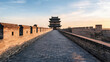 © xinxia - Ancient city wall pathway leads to a traditional multi-tiered watchtower under soft sunrise sky, showing historic Chinese fortification architecture.