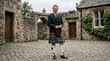 © Angavlan - Man wearing kilt in traditional Scottish attire standing outside stone buildings. The kilt features unique patterns, complemented by a blazer and accessories.