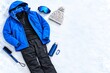 © s - 冬のウィンタースポーツ用品と防寒ウェアのフラットレイ、青いスキーウェア、ゴーグル、ニット帽、水筒、カラビナ、ブラシを雪の上に配置｜スキー・スノーボード・雪山・アウトドア