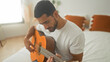 © Krakenimages.com - Man strumming acoustic guitar on bed in studio, hands fingering chords and focused expression while seated on white bedding and orange cushions; concentration.