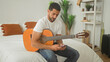 © Krakenimages.com - Man playing acoustic guitar while holding smartphone for a selfie on a bed in a residential building; relaxed creativity.