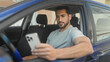 © Krakenimages.com - Man in driver seat of blue car holding smartphone with right hand and arm out the window, checking messages on screen on street; quiet concentration.