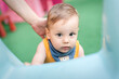 © guruXOX - A young child with light brown hair and a bib peers out from behind a colorful play structure. A hand gently guides him from behind, ensuring safety in the cheerful setting.