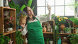 © Krakenimages.com - Woman florist in green apron holds smartphone up for selfie with potted flowers on wooden crates in a building; small business contentment.
