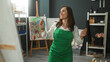 © Krakenimages.com - Woman artist in green apron holding a paintbrush to her cheek while smiling and moving in an art studio; joyful creativity.