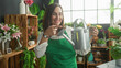 © Krakenimages.com - Woman florist in green apron holding a metal watering can and pointing with finger at its spout among potted plants in a building; care nurture joy.