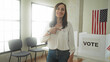 © Krakenimages.com - Woman points finger to sticker on chest at voting booth with ballot panels and american flag in polling station building; civic duty pride.