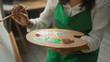 © Krakenimages.com - Woman middle age artist wearing green apron holding palette and paintbrush, hand mixing paint, paints canvas in studio; creative concentration.