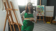 © Krakenimages.com - Woman painter holds palette and brush with hand on canvas at easel in an art studio, wearing green apron and smiling slightly; serene concentration.