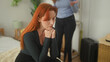 © Krakenimages.com - Woman seated on bed with hands clasped under chin and closed eyes while friend stands gesturing in bedroom; relationship strain sadness.