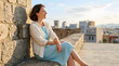 © Komang - Woman city view, travel and lifestyle at sunset for vacation, tourism and adventure. Happy person, outdoor and relaxing on stone wall with skyline, freedom and peace in town for leisure activity