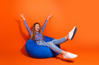 © deagreez - Joyful woman enjoying leisure time on a colorful beanbag against a bright orange background, celebrating happiness