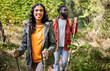 © ChaoticMind - A young Black couple enjoys a hike on a lush forest trail. The woman smiles brightly while holding trekking poles, and the man follows behind, both dressed in casual outdoor attire. Sunlight filters t
