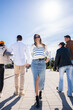 © CarlosBarquero - Vertical. Young woman walks toward the camera while friends continue ahead on a sunny promenade, creating a lively urban lifestyle and travel scene blue sky.