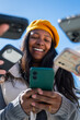 © CarlosBarquero - Vertical. Smiling Black woman looks at a smartphone while friends hold phones and cash nearby, illustrating mobile payment, digital finance and shared expenses outdoors.