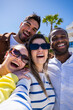© CarlosBarquero - Vertical. Four multicultural friends gather closely for a fun selfie in a modern neighborhood, smiling and enjoying leisure time together outdoors.