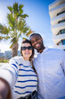 © CarlosBarquero - Vertical. Smiling interracial couple pose closely for a selfie in a bright residential area with palm trees and contemporary buildings, expressing affection and happiness.
