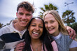 © CarlosBarquero - Three diverse young friends pose closely together and smile directly at the camera in a relaxed outdoor park setting, conveying friendship, closeness and happiness.
