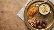© ts - Top view of a brass tray with jalebi, saffron kheer, stuffed dates, mixed nuts and a crescent star charm on a wooden table with copy space for Eid and Ramadan