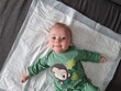 © Tina - High angle full shot of a cute happy baby lying on back on a white blanket looking at camera, infant wearing a green bodysuit with a monkey print, soft natural daytime indoor lighting