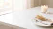 © viola - Minimalist white table setting with plate, linen napkin, wheat stalks, and a lit candle near a bright window