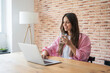 © Daniel - Woman works on a laptop at home, smiling as she types and holds a glass of water at a wooden desk. Bright window light and a brick wall create a relaxed remote office setting for freelance business.