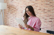 © Daniel - Young woman works on a tablet at home, reading the screen with a slight smile at a wooden desk. Casual remote work and freelance business technology concept in a bright brick-walled workspace indoors.