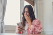 © Daniel - Woman covers her mouth while reading shocking news on a smartphone at home near a window, conveying surprise, concern, distress, bad news, and an unexpected digital notification.