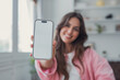 © Daniel - Woman holds a smartphone mockup with a blank white screen toward the camera, smiling warmly in a bright indoor room. Clean copy space supports app, website, advertising and interface design placement.