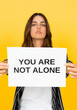 © Daniel - Woman holds a support sign reading “You are not alone” against a yellow studio background, communicating mental health awareness, compassion, reassurance, encouragement, and help for people in need.