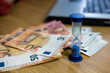 © Phoscar - Blue hourglass sitting on stack of Euro banknotes near laptop computer on wooden desk. Concept of time is money, financial investment, deadline, and deadline management