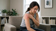© Prista - Woman experiencing shoulder pain holding her arm in discomfort seated in a living room setting