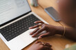 © Studio Marmellata - A Black woman works at a wooden desk, using the trackpad of her laptop. Her hands feature elegant rings and bracelets while she navigates a document on the screen.