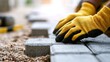 © Zarana  - Hands in yellow gloves carefully lay concrete paving stones on a gravel bed under warm sunlight. Ideal for home improvement, construction, or DIY projects.