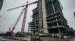© YADigital - Modern high-rise building under construction with towering red cranes, urban development scene, concrete structure, overcast sky, construction vehicles, and safety barriers