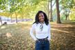 © CarlosBarquero - Young African woman smiling at the camera in a tree-lined park during autumn, wearing a scarf and white shirt. Warm backlight and soft bokeh create a relaxed, travel lifestyle mood.
