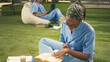 © DC Studio - Focused medical student in blue scrubs writes in an open textbook on campus grass, studying with stacked books during an outdoor learning session.